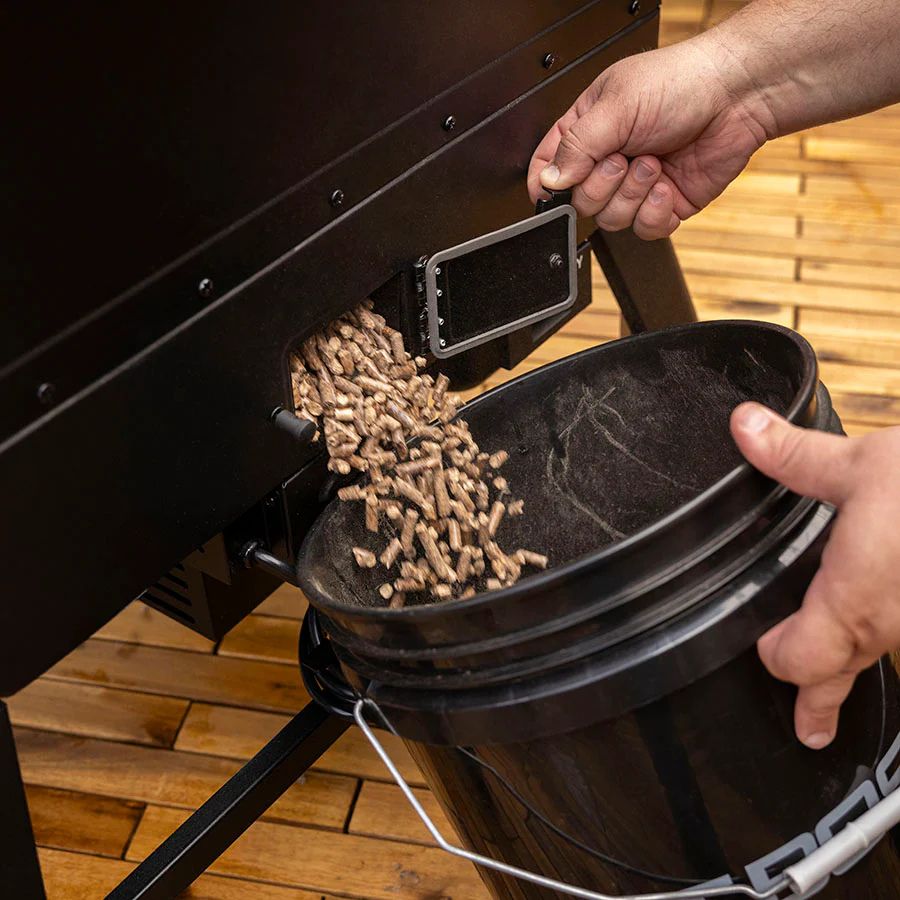 Wood pellets being emptied from the hopper of the Pit Boss Navigator 6 Series into a black bucket.