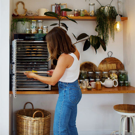 Lifestyle view of a woman using the BenchFoods 16CUDG 16-tray commercial dehydrator on a wooden countertop in a beautifully styled kitchen filled with glass storage jars of dehydrated foods.