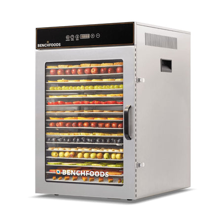 Three-quarter side angle view of a BenchFoods 16CUDG 16-tray commercial food dehydrator fully loaded with vibrant rows of sliced apples, grapes, kiwis, and citrus fruit behind a glass door.