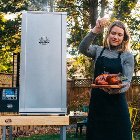 Woman seasoning smoked turkey next to Bradley Digital 6-Rack Electric Smoker outdoors