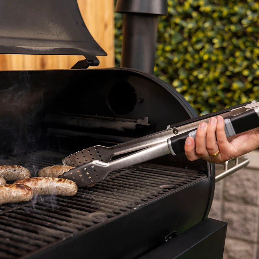 A hand holding Pit Boss BBQ tongs to turn sausages on a smoking grill