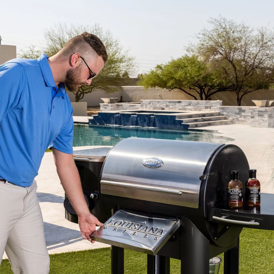 Man using Louisiana Grills Founders Legacy 800 Outdoor Pellet Grill in a backyard setting, with BBQ sauces on the side table, highlighting outdoor cooking experience.