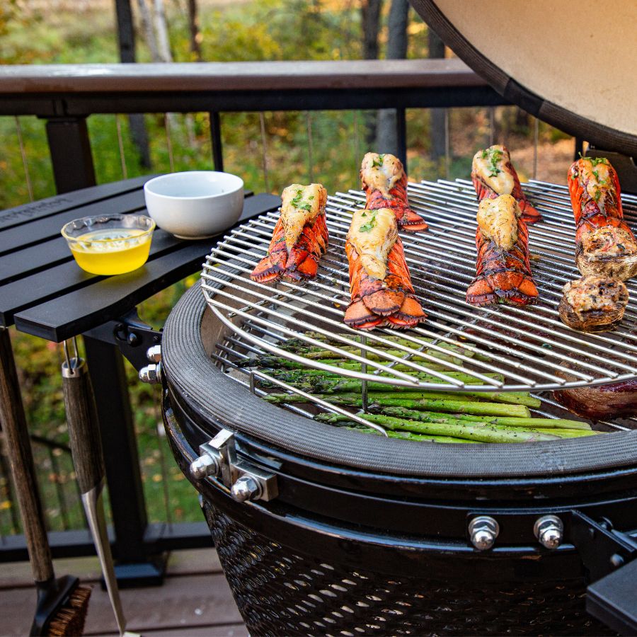 Louisiana Grills 24" Ceramic Kamado Charcoal Grill with lobster tails, asparagus, and steak cooking on the grill grates. A bowl of melted butter sits nearby for basting.