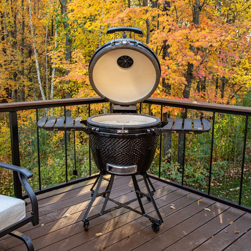 Louisiana Grills 24" Ceramic Kamado Charcoal Grill set up on a wooden deck surrounded by autumn trees, perfect for outdoor cooking and smoking.