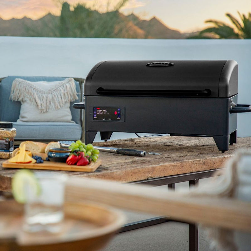 Louisiana Grills 300 Black Label Portable Wood Pellet Grill on a rustic outdoor table with fresh food, set against a scenic backdrop, ideal for backyard grilling.