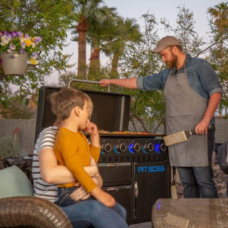 Person grilling on the Pit Boss 5-Burner Ultimate Griddle in a backyard at dusk, with family nearby