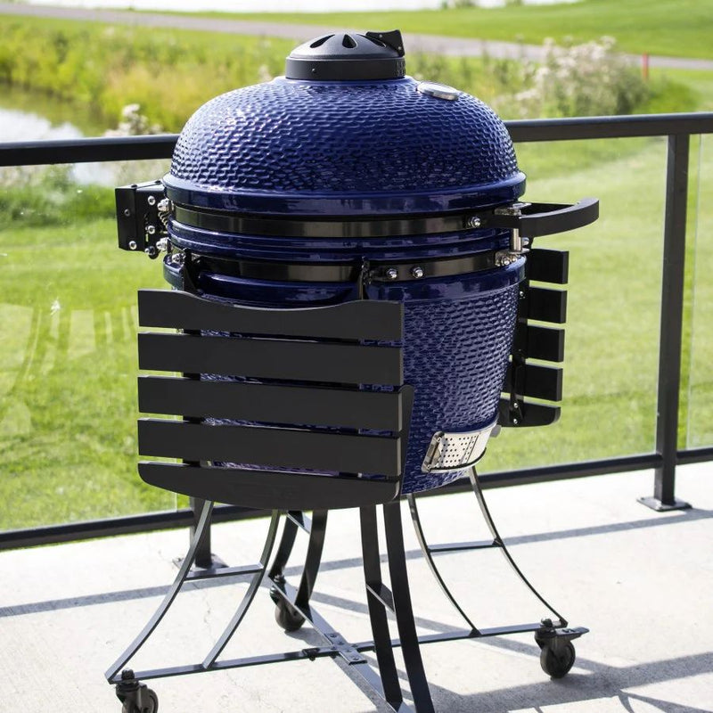 The Pit Boss K24 Ceramic Charcoal Grill set up on an outdoor patio, showcasing its premium blue ceramic design and spacious side shelves for food prep.